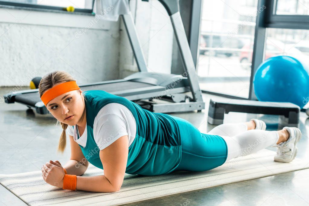attractive overweight woman doing plank exercise on fitness mat near treadmill