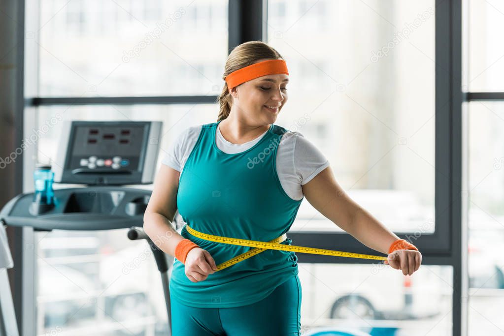 cheerful plus size girl looking at measuring tape while measuring waist in gym