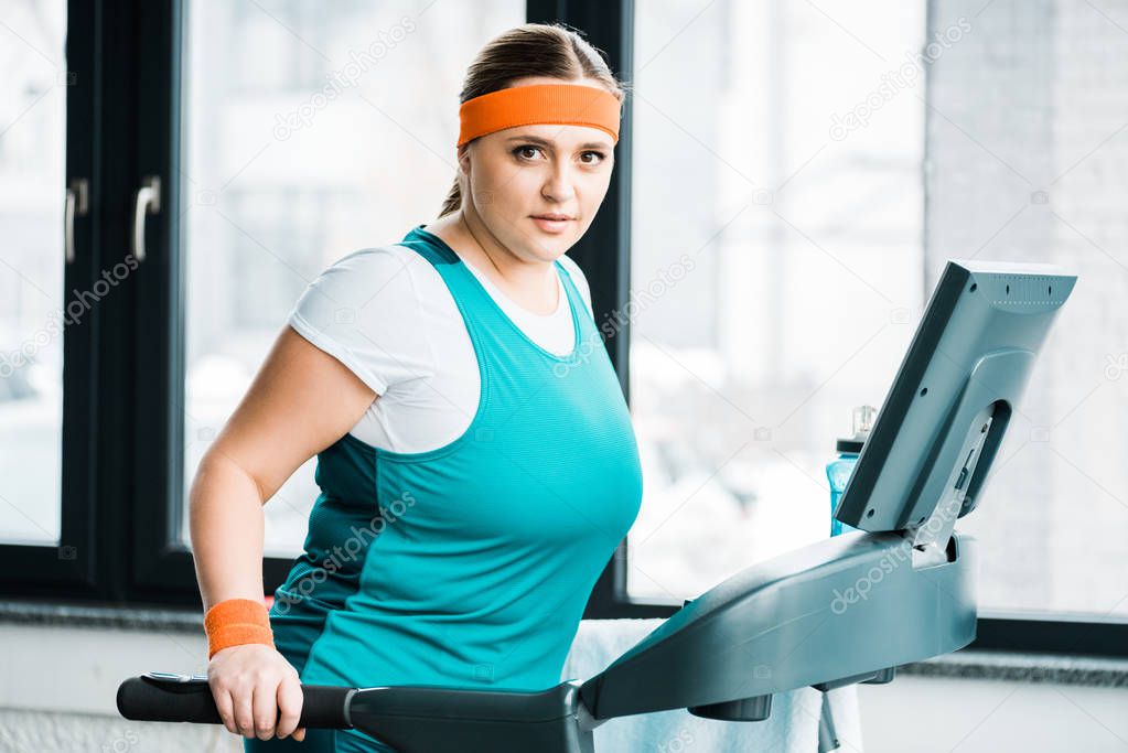 attractive overweight girl exercising on treadmill while looking at camera in gym