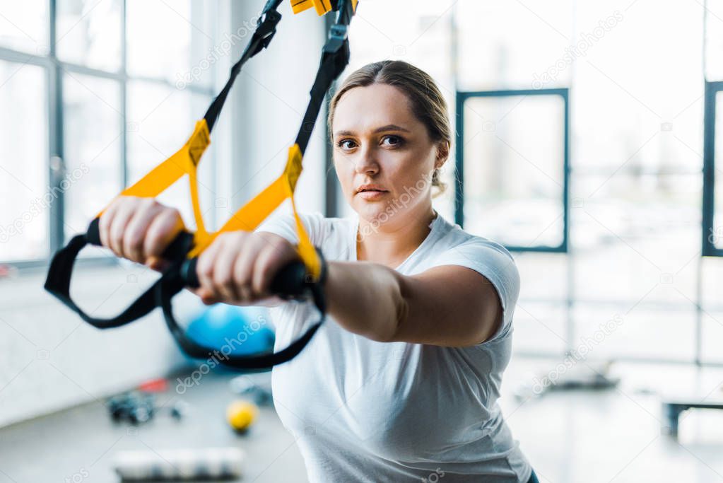 confident overweight woman training arms with suspension straps in gym
