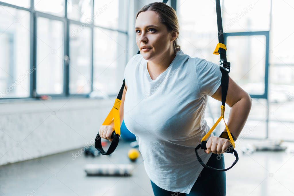 focused overweight girl training arms with suspension straps in gym
