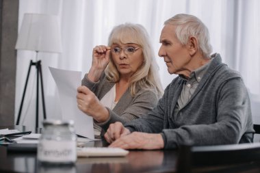 surprised senior couple sitting at table and looking at bills  clipart