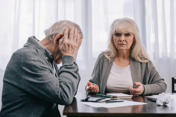 upset senior man with hands on head sitting at table with woman looking at camera