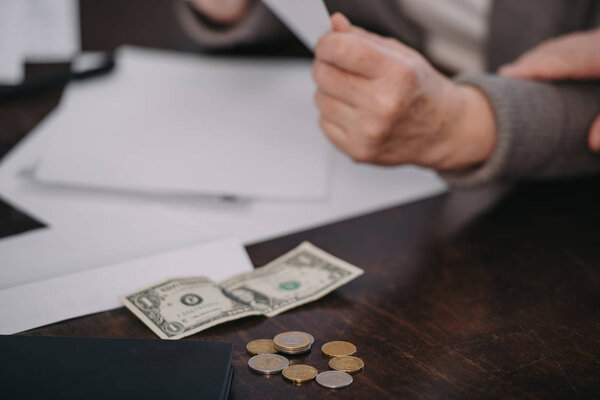 cropped view of senior woman sitting at table with paperwork, coins and banknote