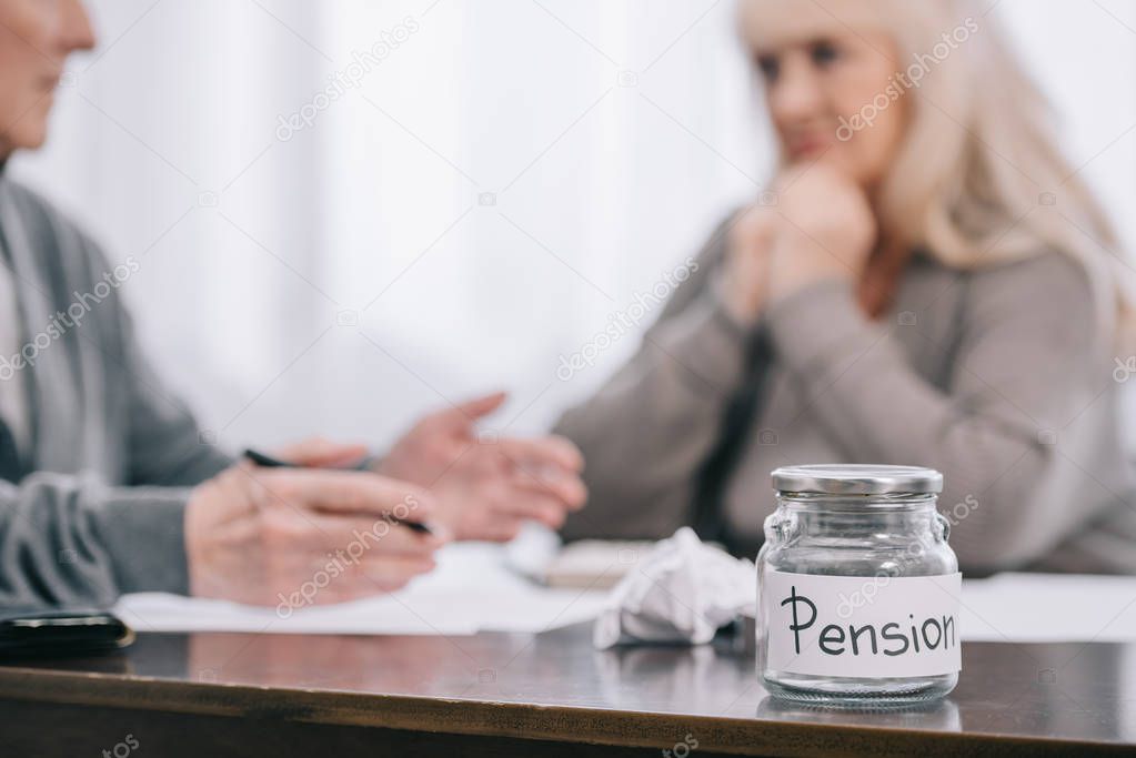 selective focus of 'pension' word on empty glass jar with senior couple sitting at table on background