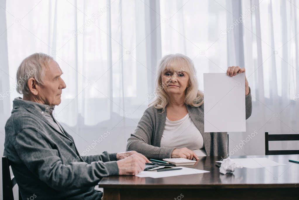 senior woman holding empty paper and looking at camera while sitting at table with man