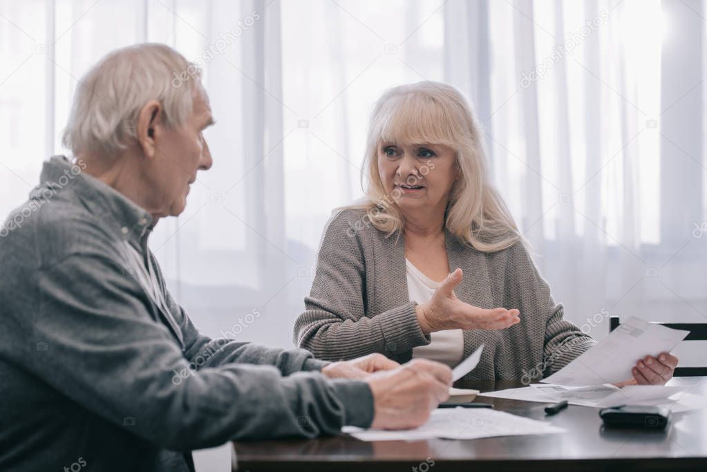 senior couple in casual clothes sitting at table and holding bills 