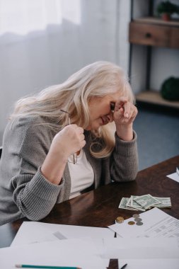 tired senior woman sitting at table and having headache while counting money at home clipart