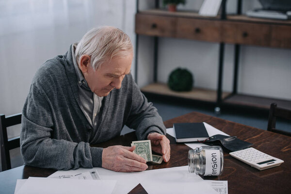 senior man sitting at table with money, paperwork and glass jar with 'pension' lettering