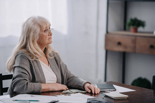 старшая женщина сидит за столом с деньгами и бумажной работой во время подсчета бюджета на дому
 