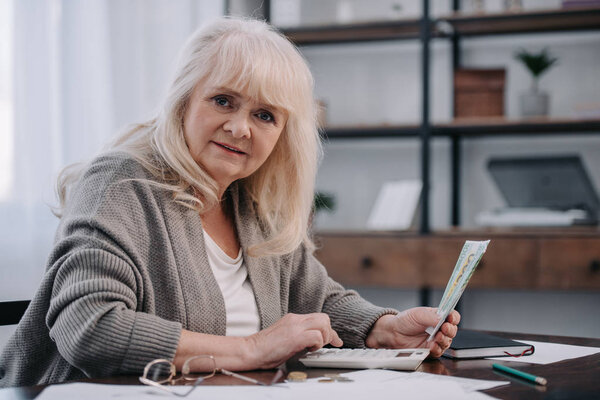 старшая женщина сидит за столом, смотрит в камеру, использует калькулятор и подсчитывает деньги
