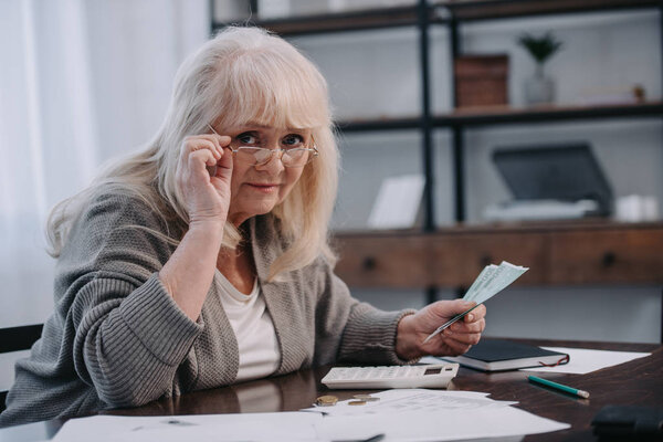 senior woman sitting at table with paperwork, looking at camera and counting money