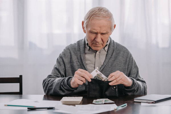 male pensioner sitting at table with paperwork and putting money in wallet 