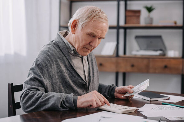 senior man sitting at table with paperwork and using calculator while counting money 