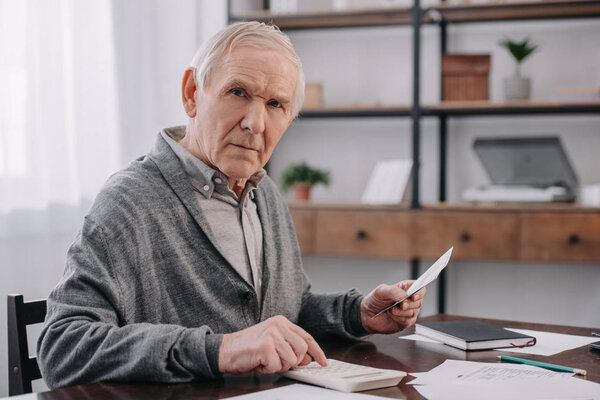 senior man sitting at table with paperwork, looking at camera and using calculator while counting money 