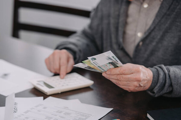 cropped view of senior man sitting at table with paperwork and using calculator while counting money 