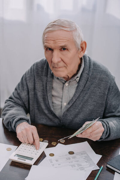 senior man sitting at table with paperwork, using calculator while counting money and looking at camera