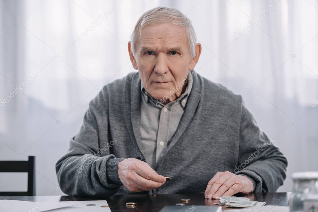senior man sitting at table, looking at camera and counting money at home