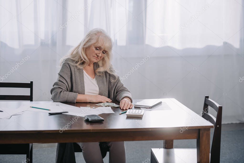 senior woman sitting at table with money and paperwork while counting budget at home with copy space