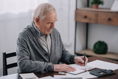 senior man sitting at table with paperwork and writing in notebook clipart