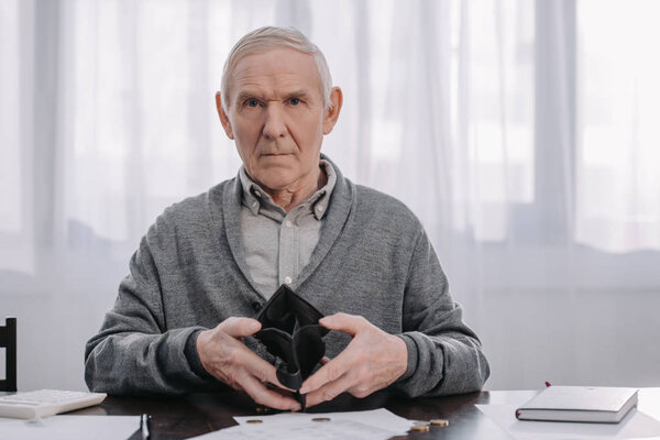 male pensioner sitting at table with paperwork, looking at camera and holding empty wallet 
