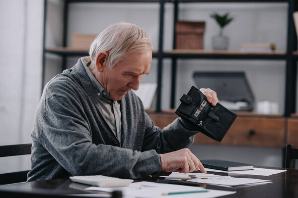 senior man sitting at table with paperwork, holding wallet and counting money at home