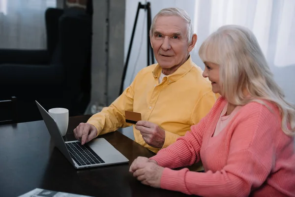 高级夫妇坐在沙发上与笔记本电脑和信用卡 而在家里做网上购物 — 图库照片