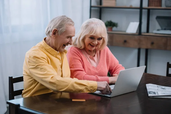 微笑的老夫妇使用笔记本电脑 而坐在家里的桌子 — 图库照片
