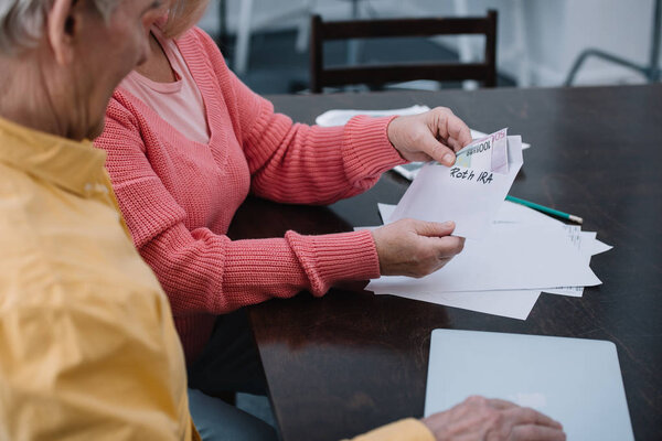 partial view of senior couple holding envelope with 'roth ira' lettering and money while sitting at table