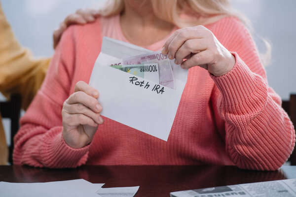 partial view of woman holding envelope with 'roth ira' lettering and money