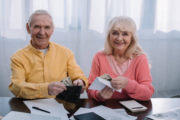 пожилая пара держит бумажник и конверт с деньгами, глядя в камеру
