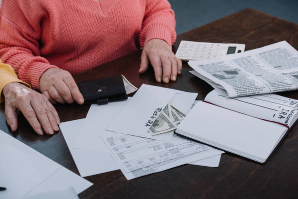 cropped view of senior couple sitting at table with paperwork, envelope with 'roth ira' lettering, money and notebook