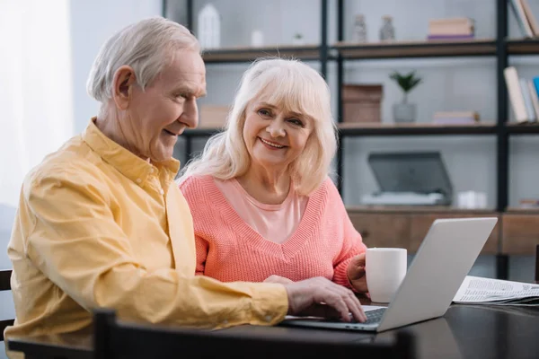 微笑的老夫妇在五颜六色的衣服坐在桌子上 并使用笔记本电脑 — 图库照片