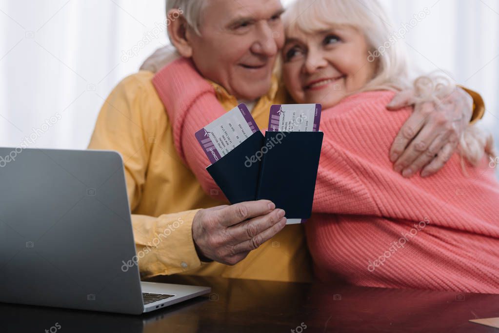 happy senior couple sitting at table with laptop, hugging and holding air tickets with passports at home 