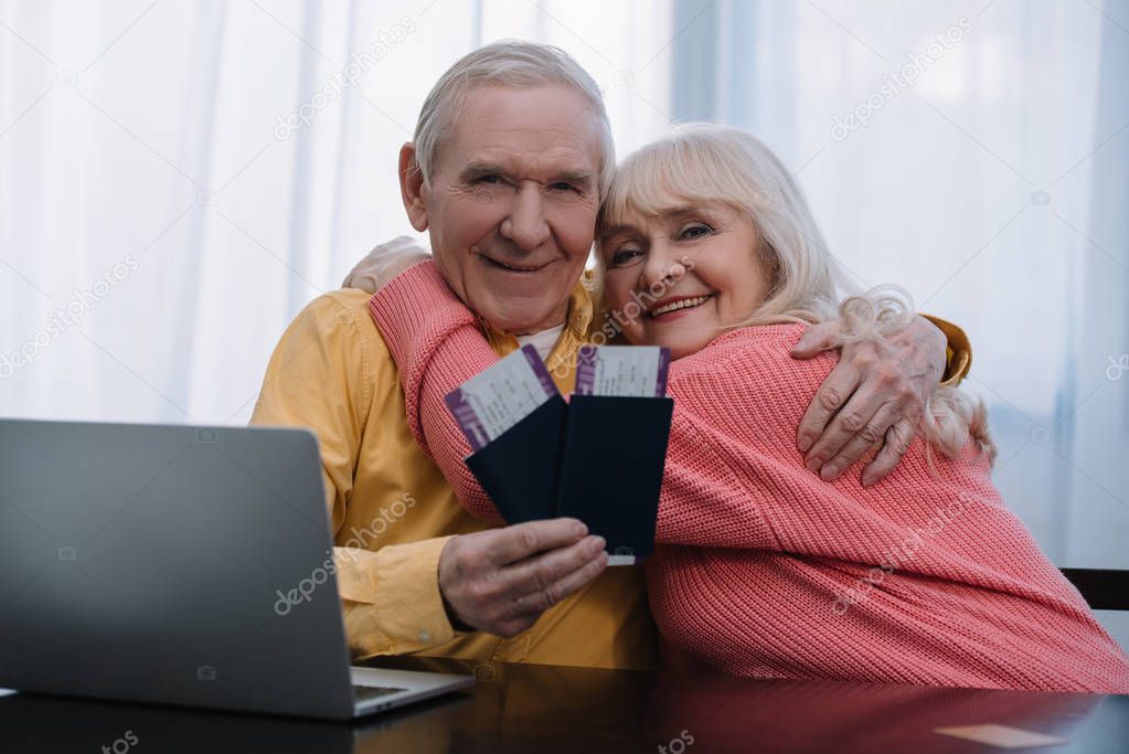 happy senior couple sitting at table with laptop, hugging and holding air tickets with passports at home 