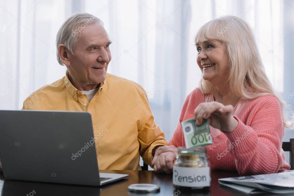 senior couple sitting at table with laptop while woman putting money in glass jar with 'pension' lettering