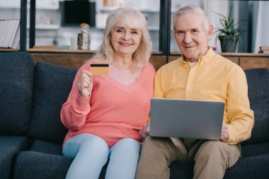 laptop ve kredi kartı ile kanepede oturan ve evde online alışveriş yaparken kameraya bakarak mutlu üst düzey Çift