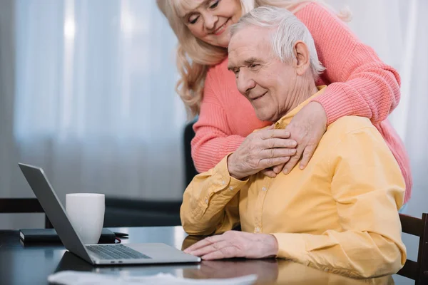 Sorrindo Casal Sênior Abraçando Enquanto Homem Sentado Mesa Usando Laptop — Fotografia de Stock