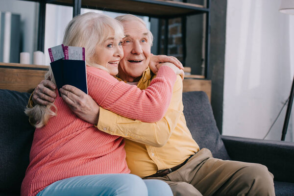 happy senior couple hugging and holding air tickets with passports at home 