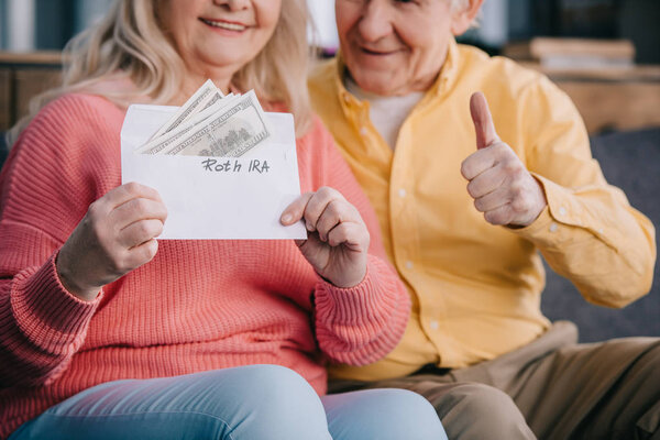 cropped view of senior couple showing thumb up sign while holding envelope with 'roth ira' lettering and dollar banknotes