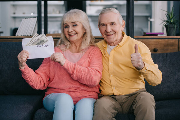 senior couple showing thumb up sign while holding envelope with 'roth ira' lettering and dollar banknotes