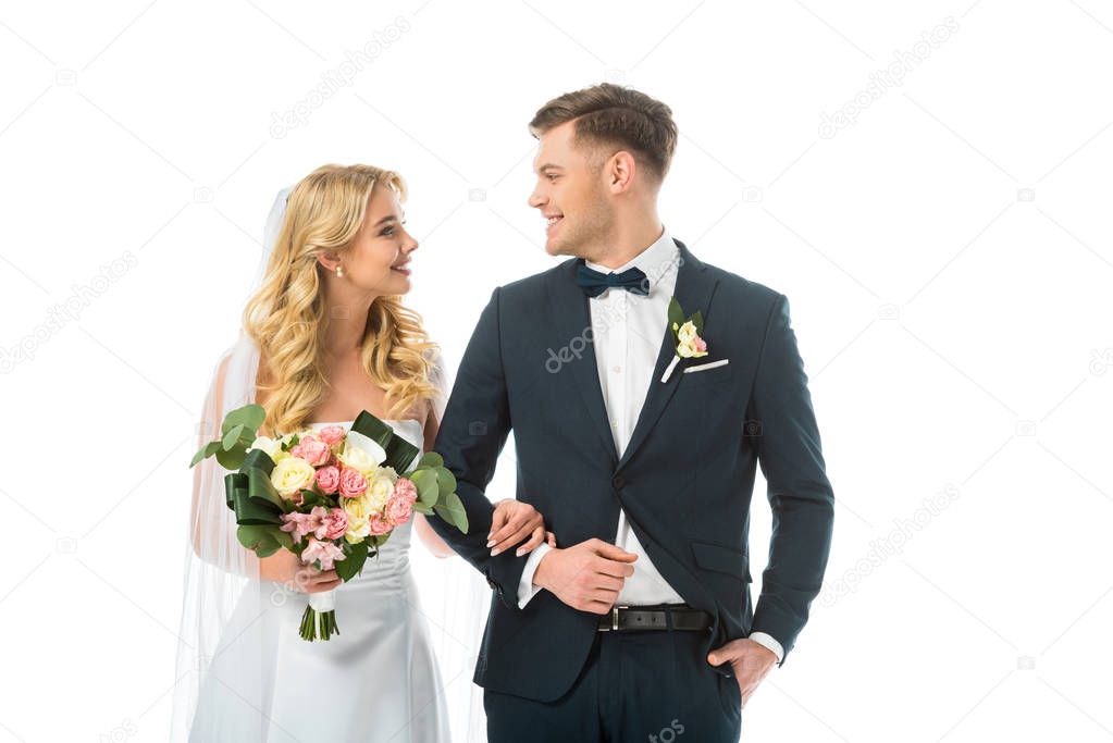 smiling bride with wedding bouquet, and happy groom in black suit looking at each other isolated on white