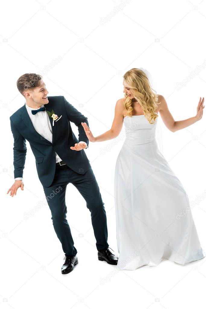 cheerful groom in black elegant suit dancing with happy bride in white wedding dress isolated on white
