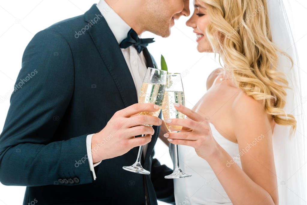 happy bride and groom clinking glasses of champagne while standing face to face isolated on white 