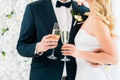 abgeschnittene Ansicht des Bräutigams und der Braut mit Champagnergläsern auf weißem Blumenhintergrund