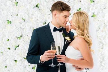 mutlu genç damat ve gelin yüz yüze ve elinde bardak şampanya beyaz çiçek arka planda duran