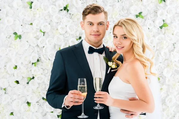 快乐的新郎和新娘拿着香槟眼镜 看着相机在白色花卉背景 — 图库照片