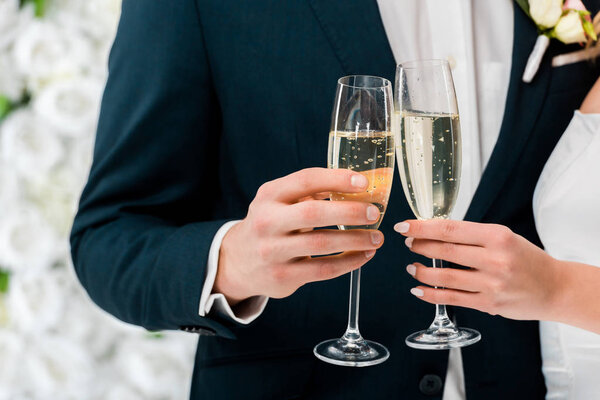 partial view of groom and bride with glasses of champagne on white floral background