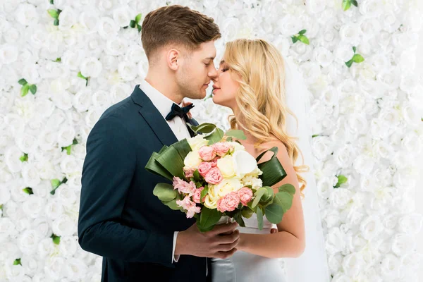 Feliz Jovem Noiva Noivo Beijando Enquanto Segurando Buquê Casamento Fundo — Fotografia de Stock