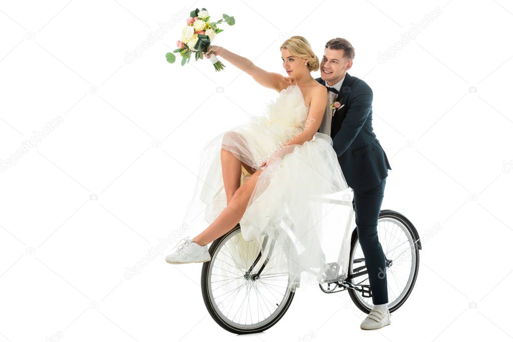 beautiful bride holding wedding bouquet in raised hand while sitting on bicycle together with groom isolated on white